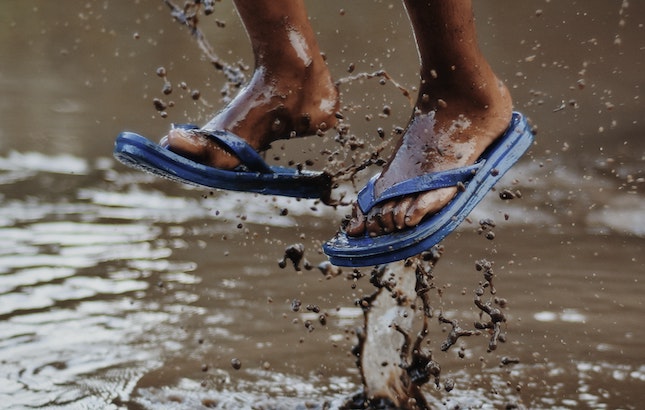 Two feet in flip-flops jumping over muddy water with muddy droplets splashing.