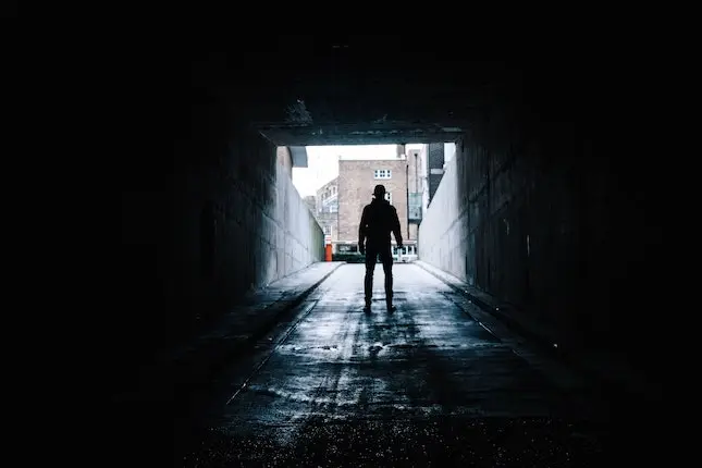 man walking out of a dark tunnel into an unknown area with buildings